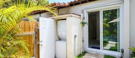 Una casa pequeña de una planta con techo de tejas, una puerta principal, una puerta corredera de vidrio, una lavadora y una valla de madera.