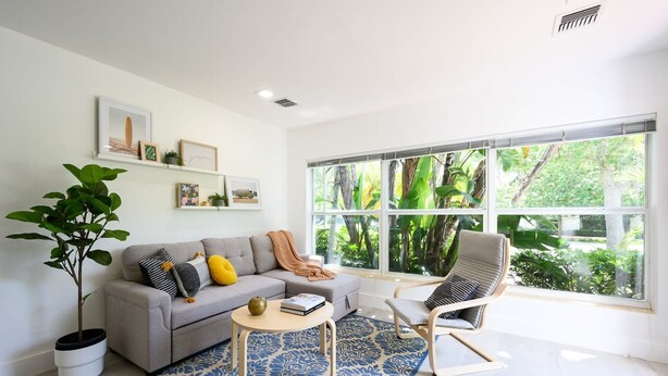Un salon moderne avec un canapé gris, un tapis bleu à motifs, une table basse en bois et un fauteuil. Une grande fenêtre offre une vue sur la verdure à l’extérieur.
