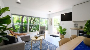 Un salon moderne comprenant un canapé, une table basse et un coin repas avec une table et des chaises. Une grande fenêtre offre une vue sur la verdure environnante.