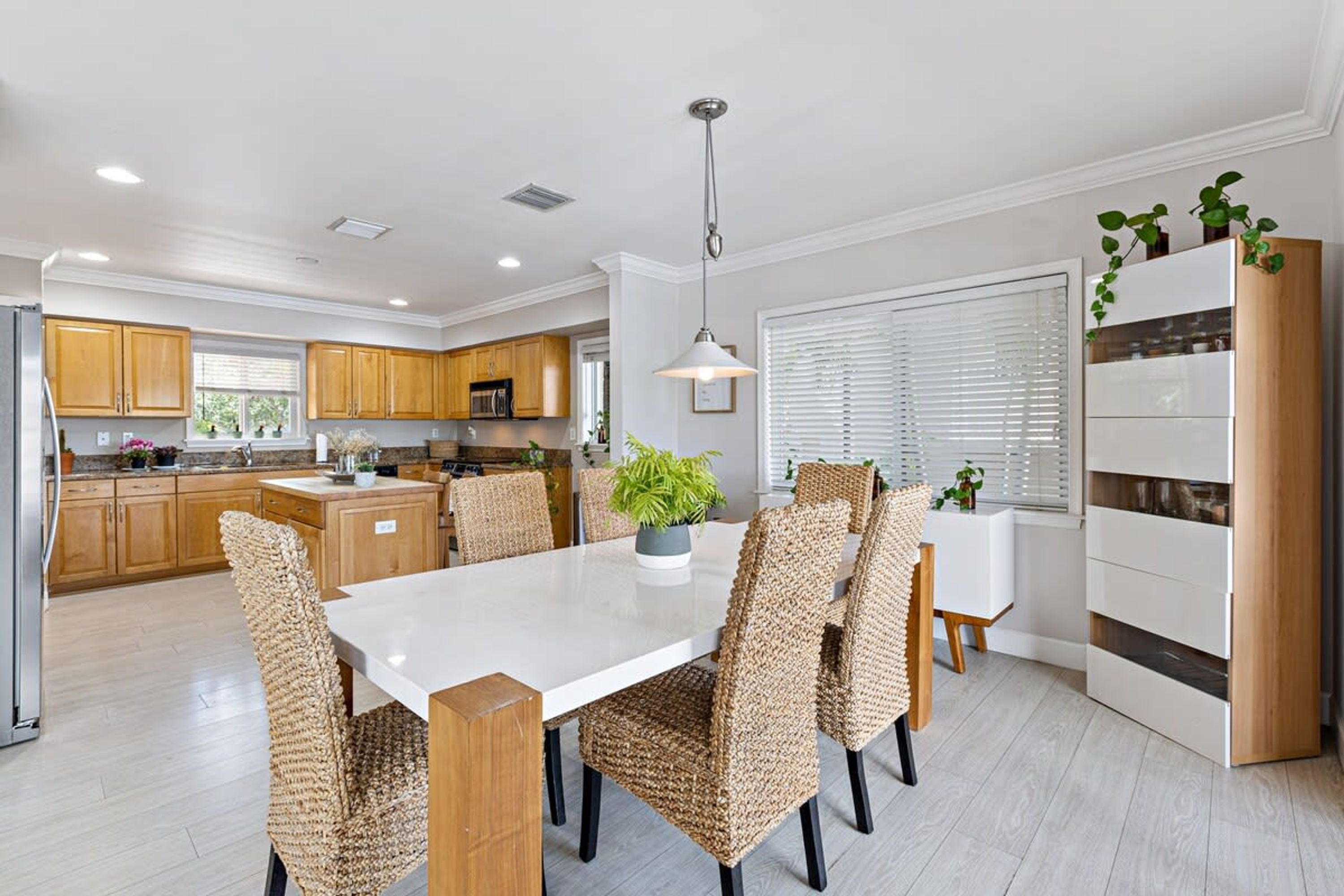 A modern kitchen with wooden cabinets, a white dining table, woven chairs, and a pendant light.