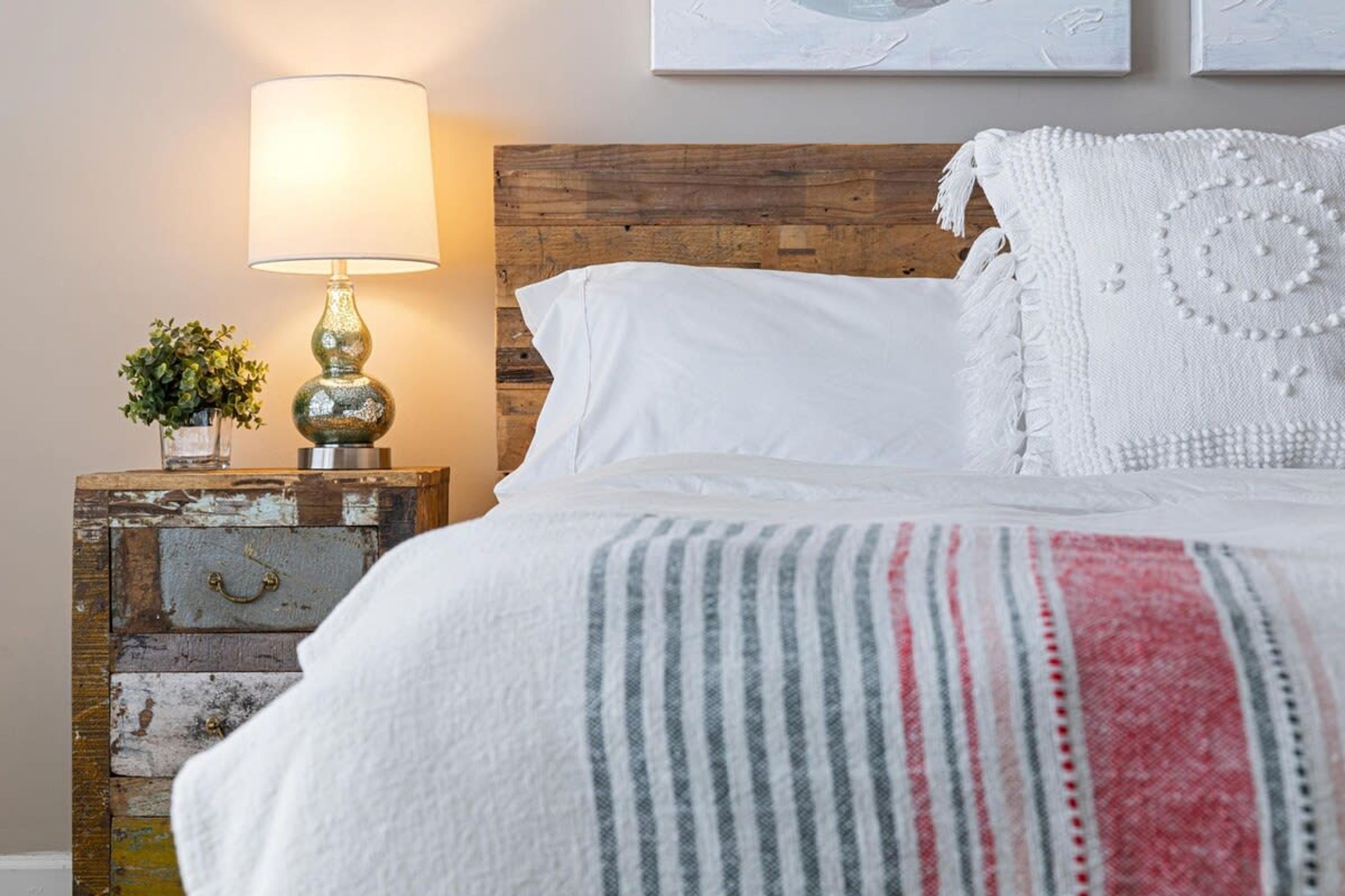 A neatly made bed with white linens, a striped blanket, and decorative pillows. A vintage lamp and a small plant are on a wooden nightstand beside the bed.