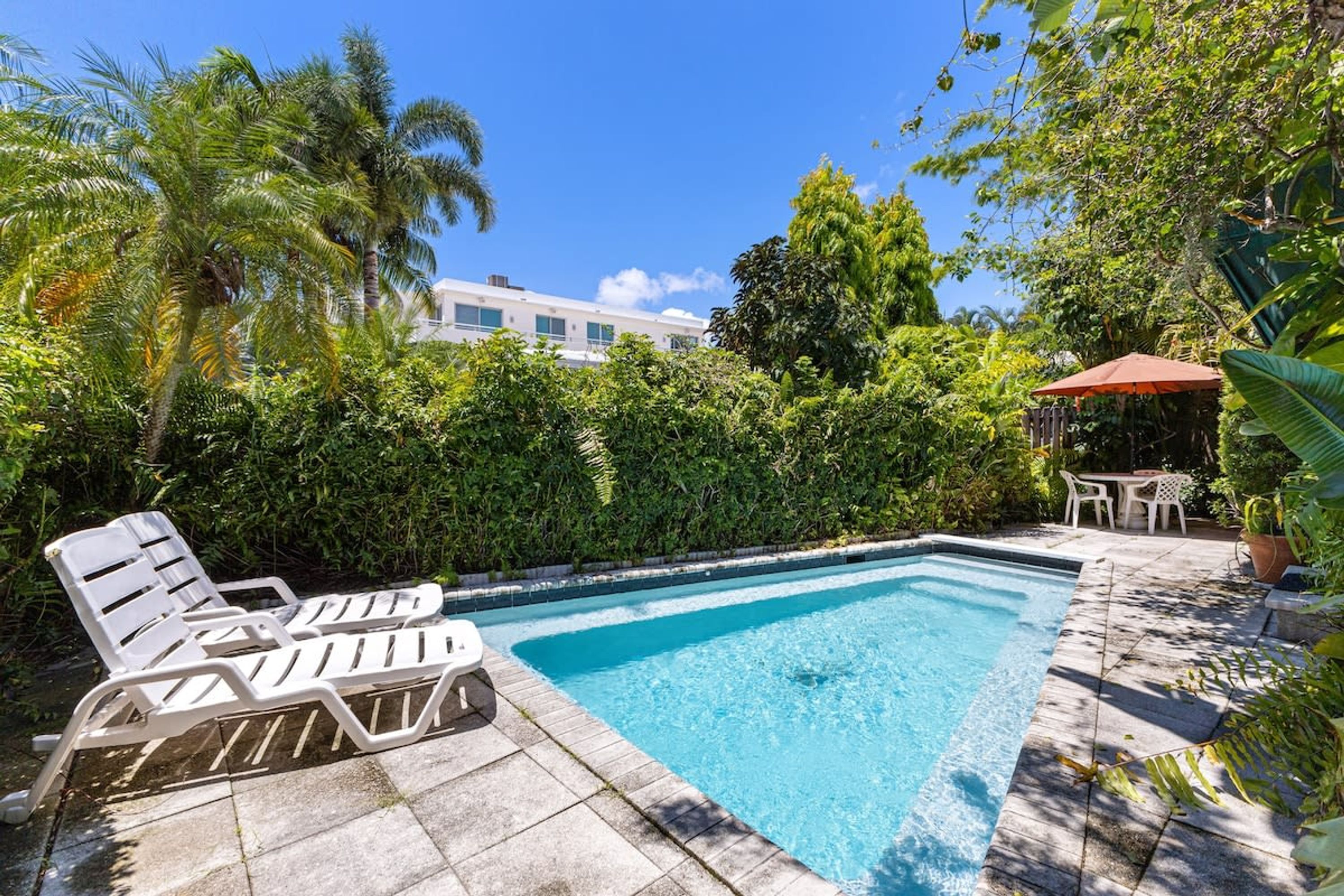 A swimming pool with lounge chairs, surrounded by lush greenery and a clear blue sky.