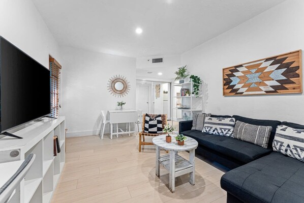 A modern living room with a flat-screen TV, a sofa, a coffee table, and a dining area with a white table and chairs.