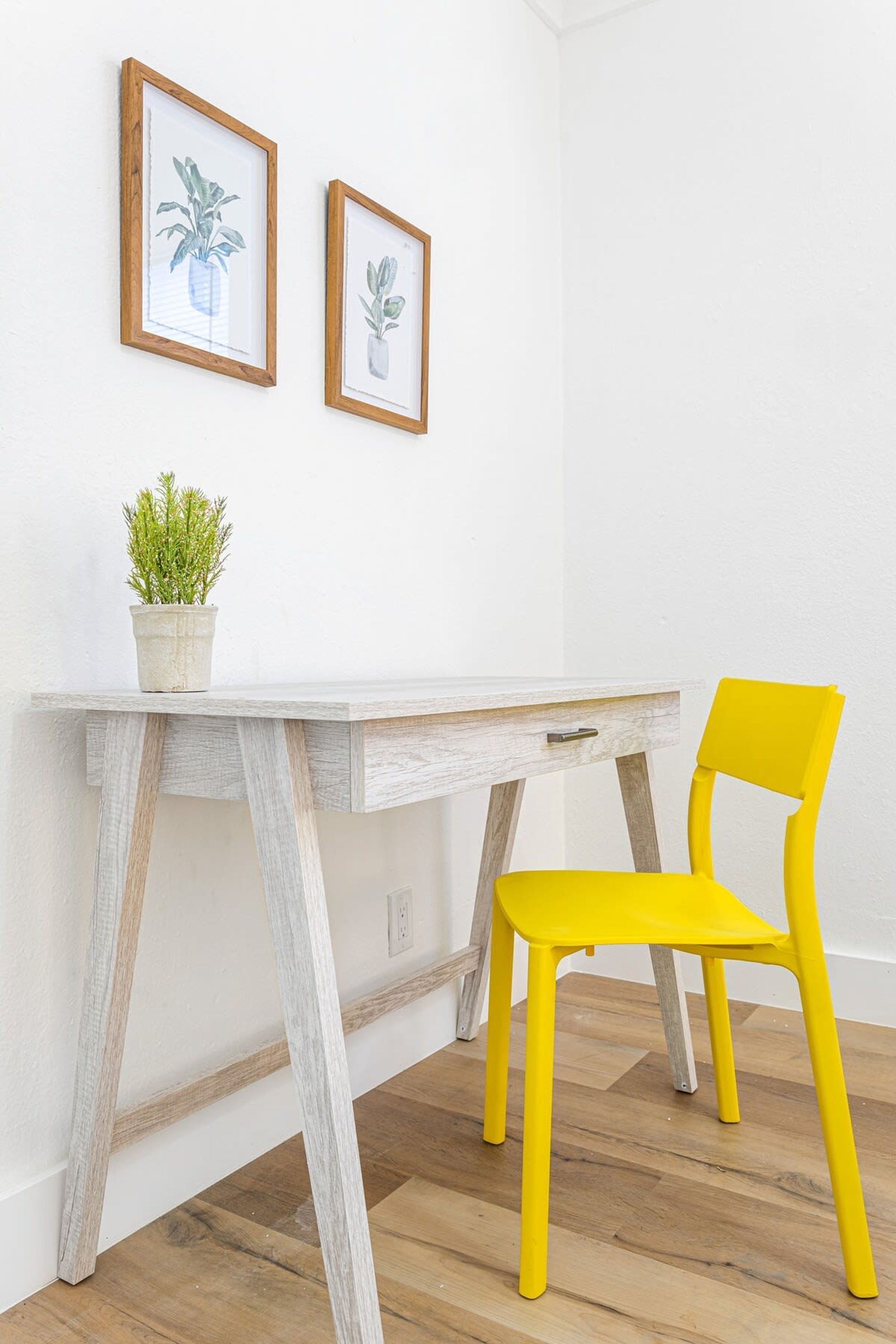 Un bureau blanc avec un cadre en bois, une chaise jaune et deux reproductions botaniques encadrées accrochées au mur.