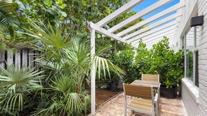 A covered patio with a wooden table and chairs, surrounded by various potted plants and trees.