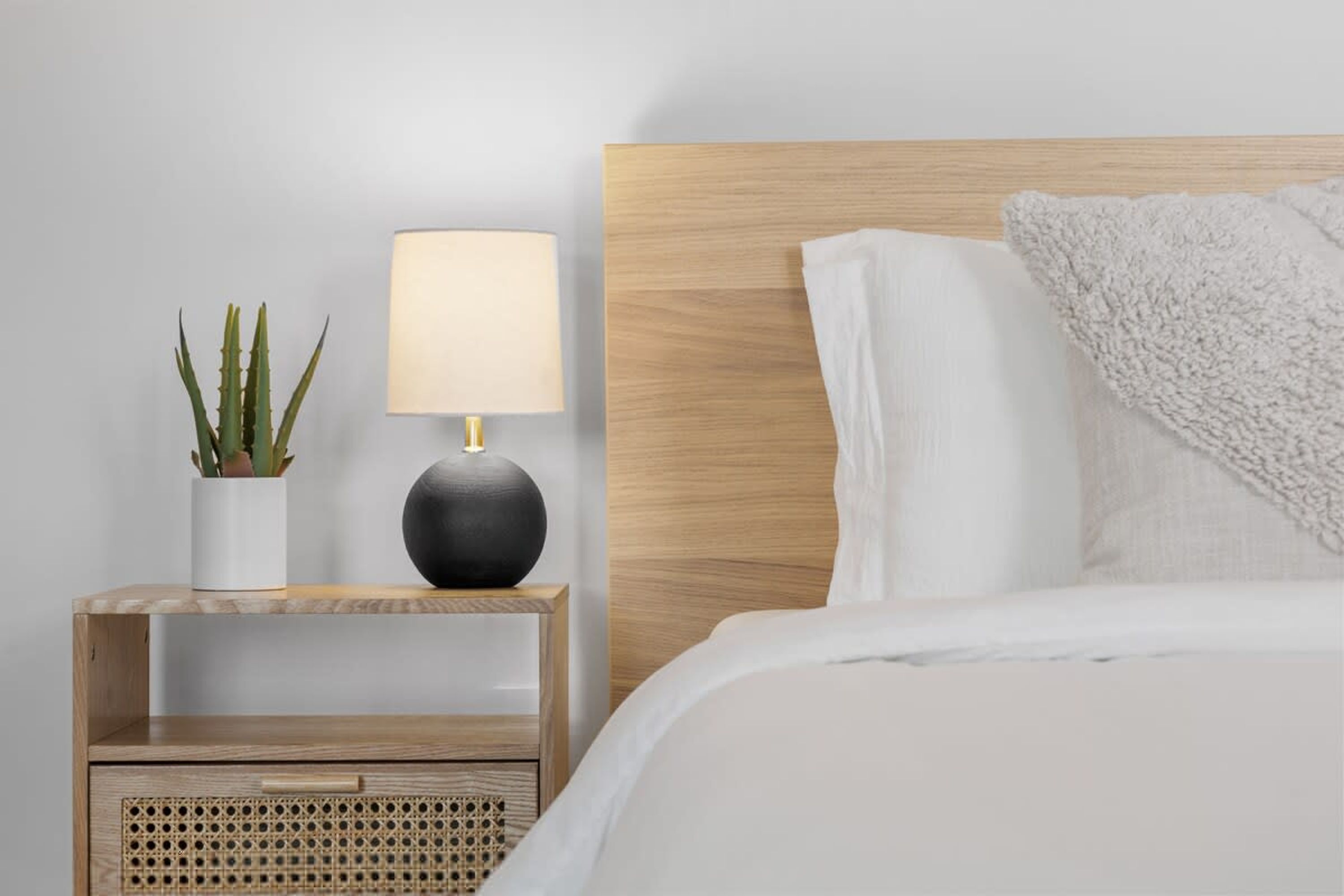 Un lit bien fait, avec une couette blanche et des oreillers, une lampe de chevet, une petite table de nuit en bois dotée d’un tiroir en rotin et un pot contenant une plante d’aloès.