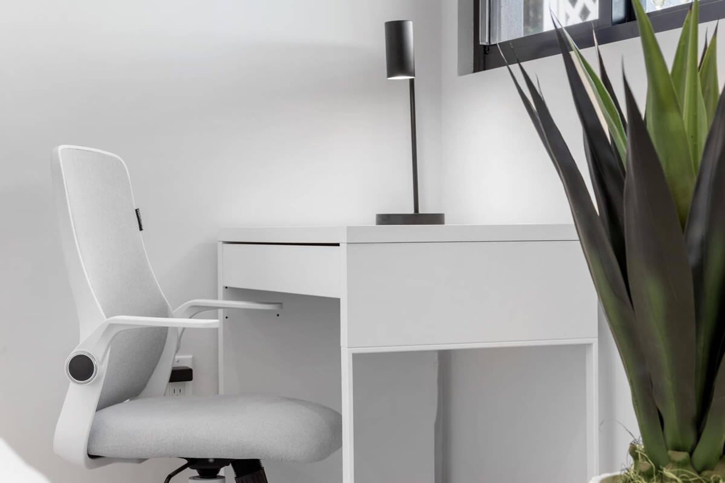 Una silla de oficina moderna, un escritorio blanco, una lámpara de pie negra y una planta en maceta.