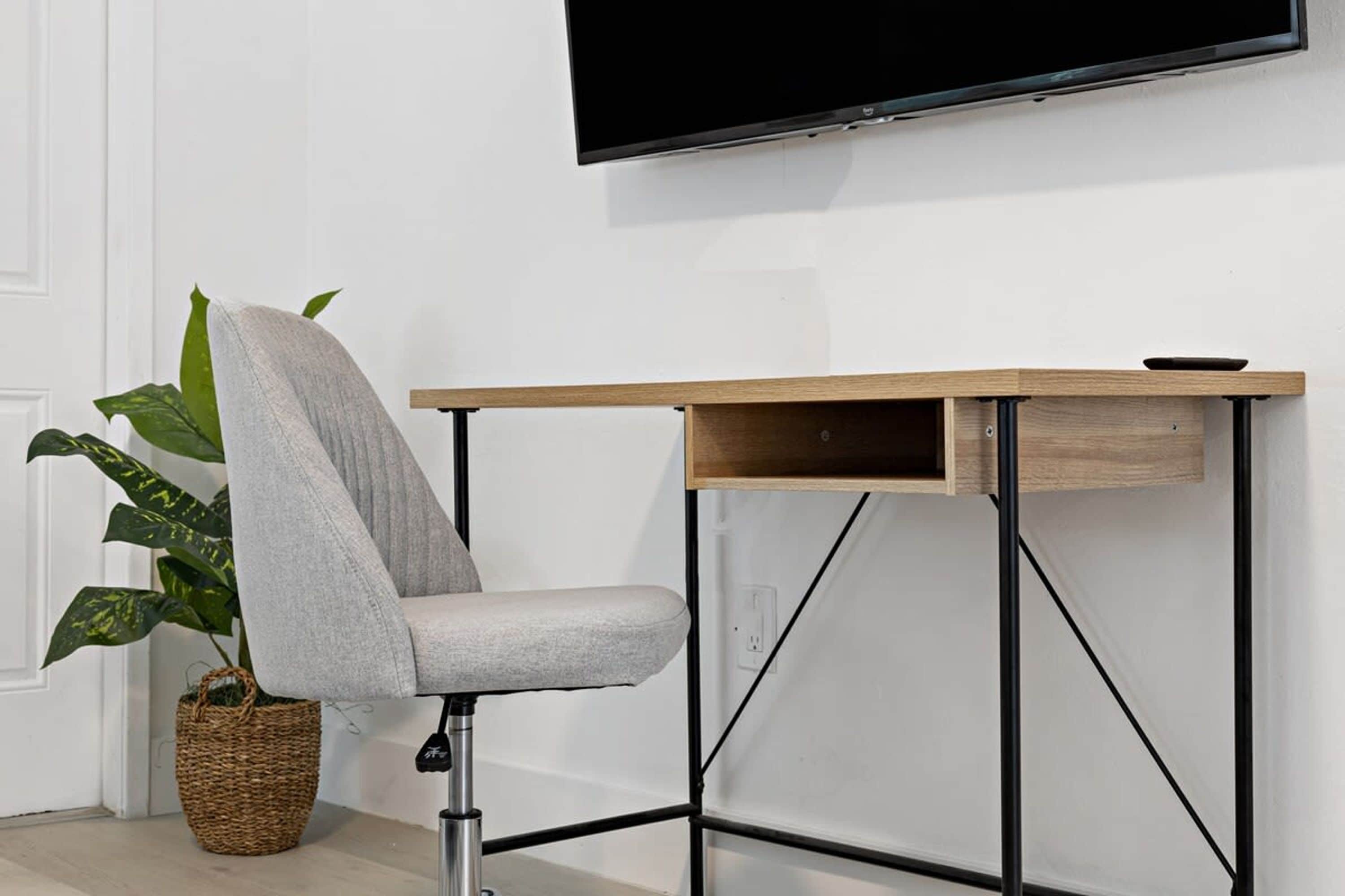 Moderni toimistotuoli, jossa on harmaa istuintyyny, puinen työpöytä, jonka runko on mustaa metallia, sekä suuri, seinälle asennettu litteänäyttöinen televisio.