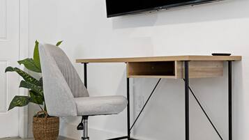 Moderni toimistotuoli, jossa on harmaa istuintyyny, puinen työpöytä, jonka runko on mustaa metallia, sekä suuri, seinälle asennettu litteänäyttöinen televisio.