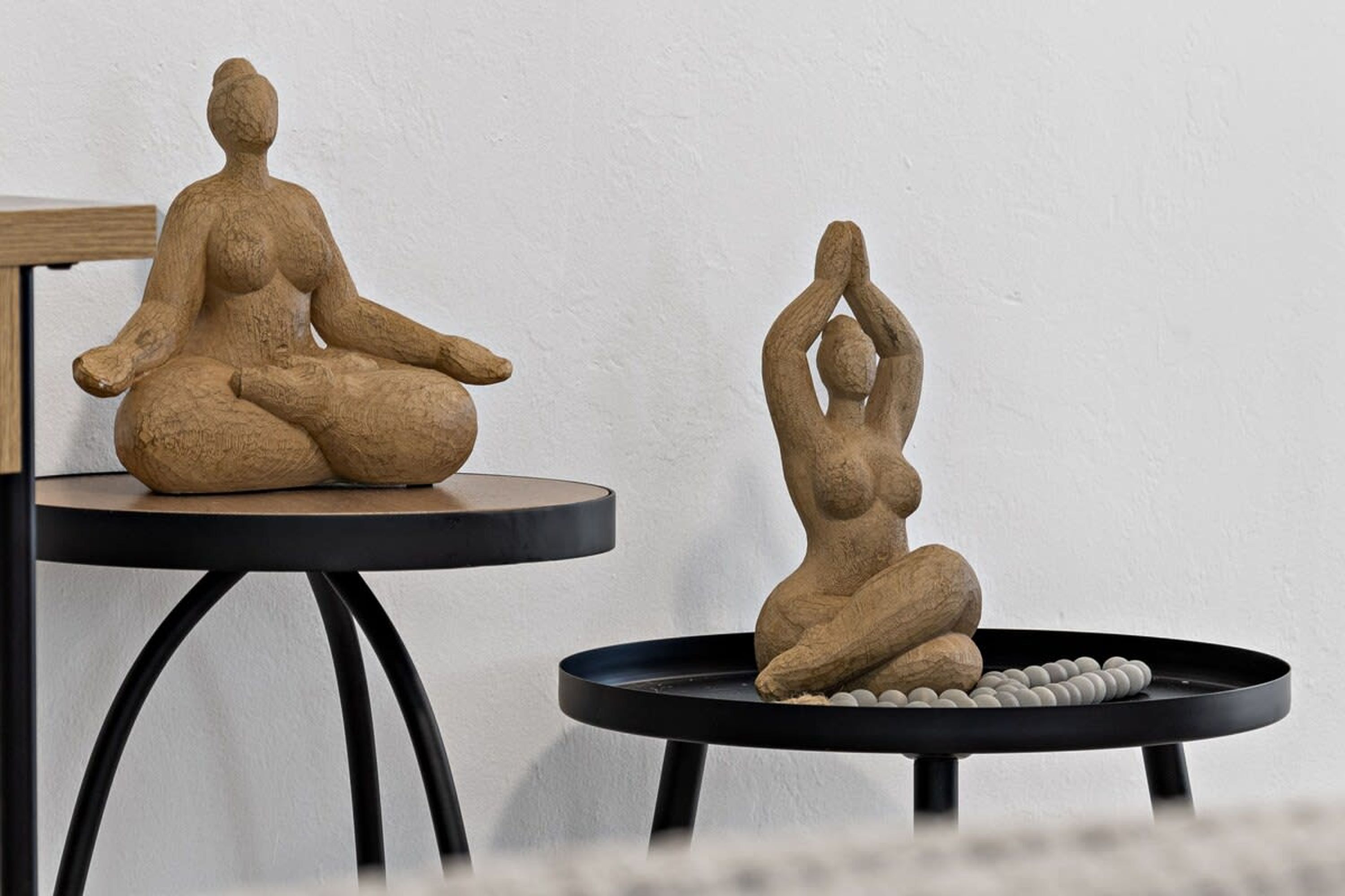 Due sculture in legno su tavoli neri, con una parete neutra sullo sfondo.
