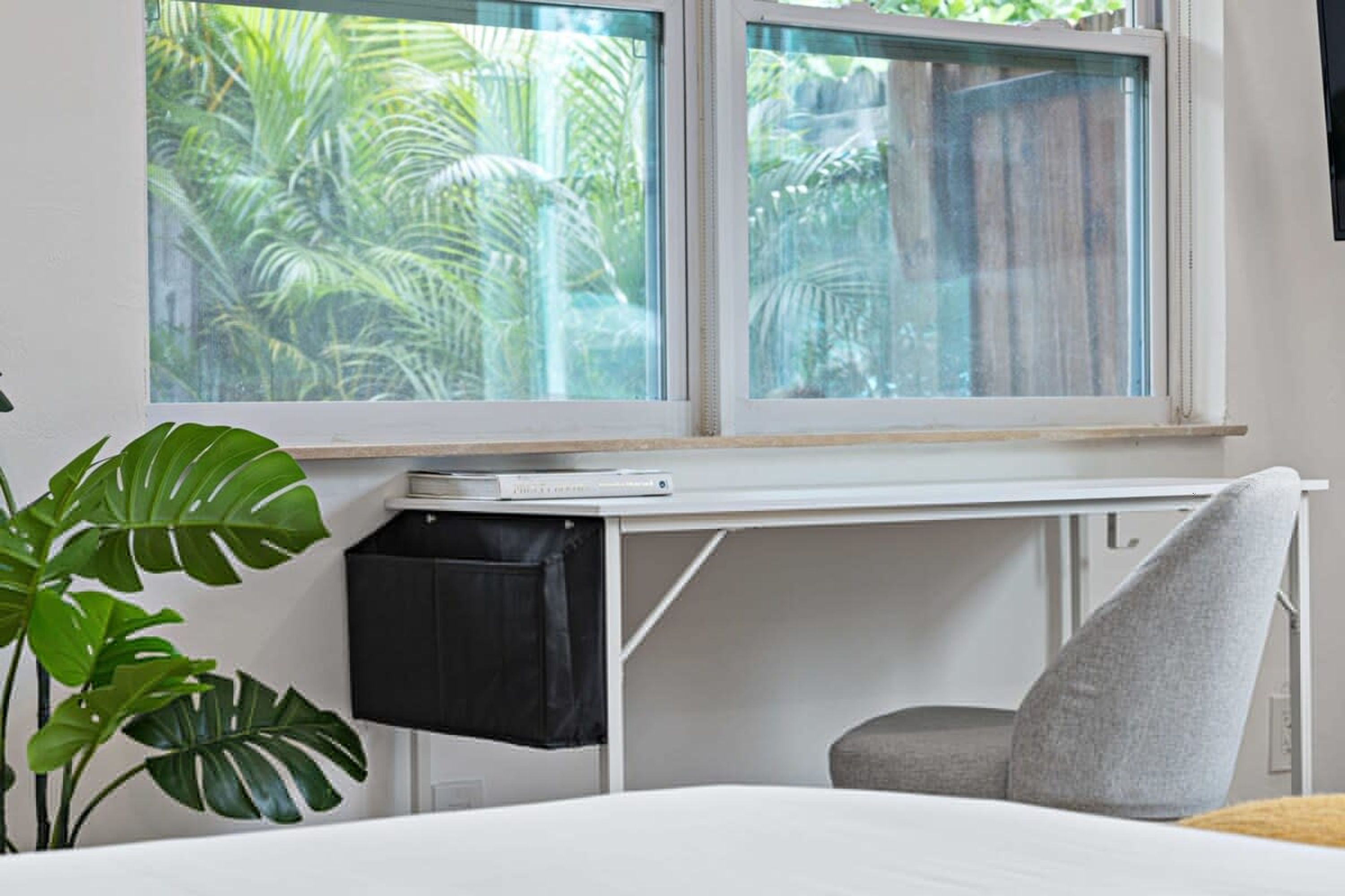 A bedroom with a bed, a desk, and a chair. There are two windows with a view of greenery outside.