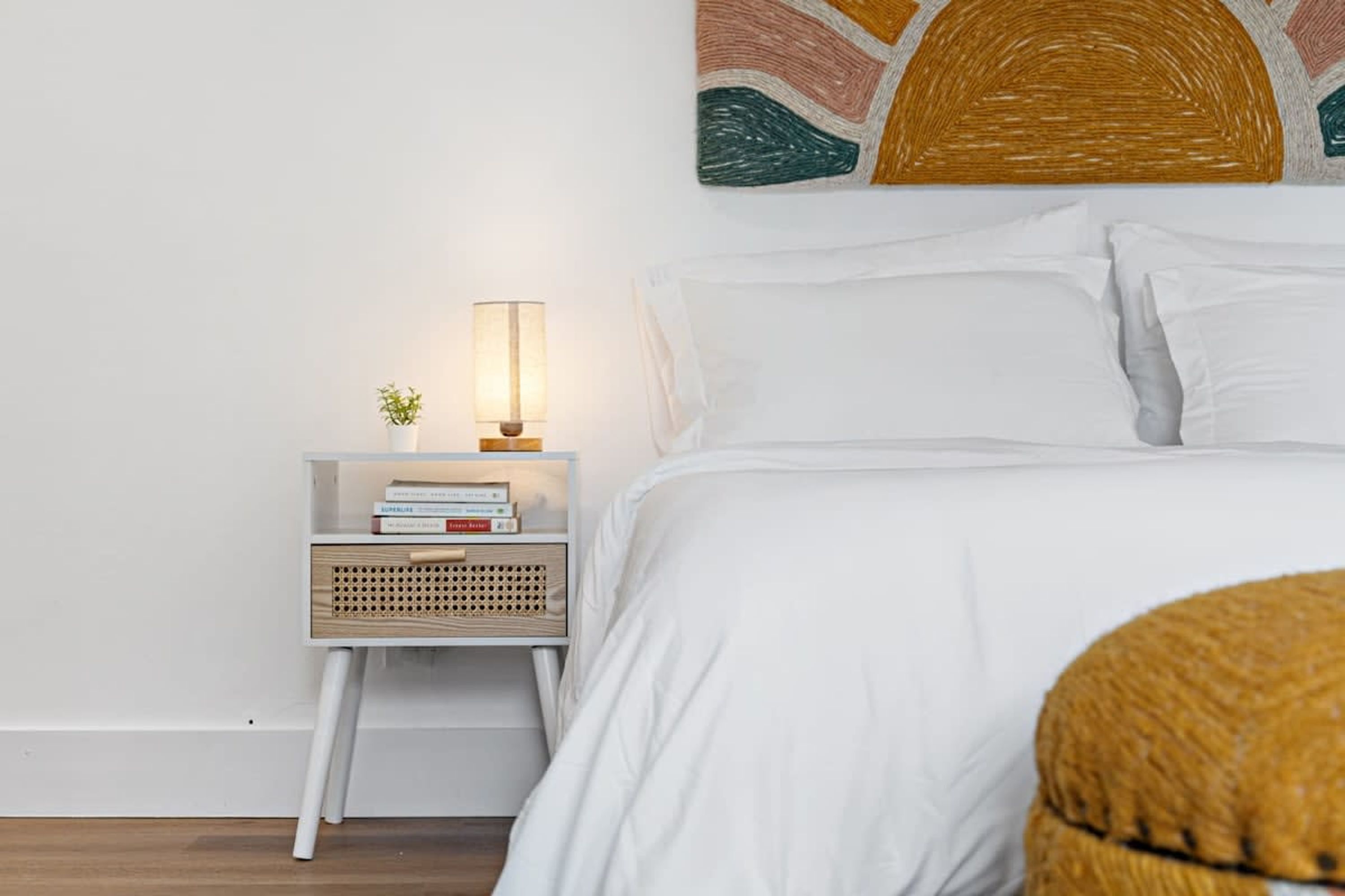 A neatly made bed with white linens, a bedside lamp, and a small wooden side table with a plant and books.