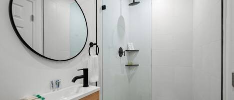 Un baño moderno con un espejo redondo, un lavamanos blanco y una zona de ducha con mampara de vidrio.