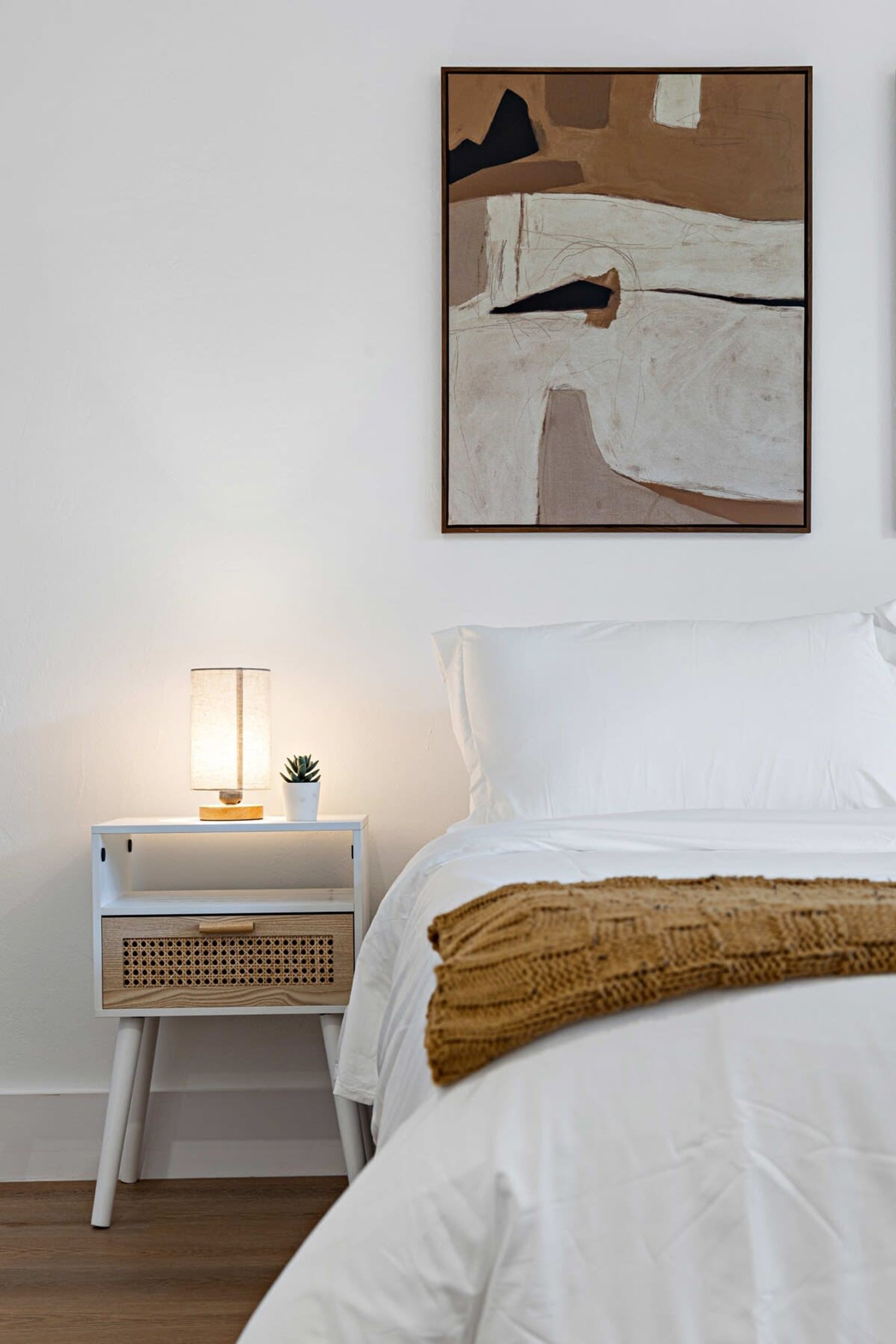Ein Schlafzimmer mit einem weißen Bett, einem Nachttisch mit Lampe und einem an der Wand befestigten abstrakten Gemälde.