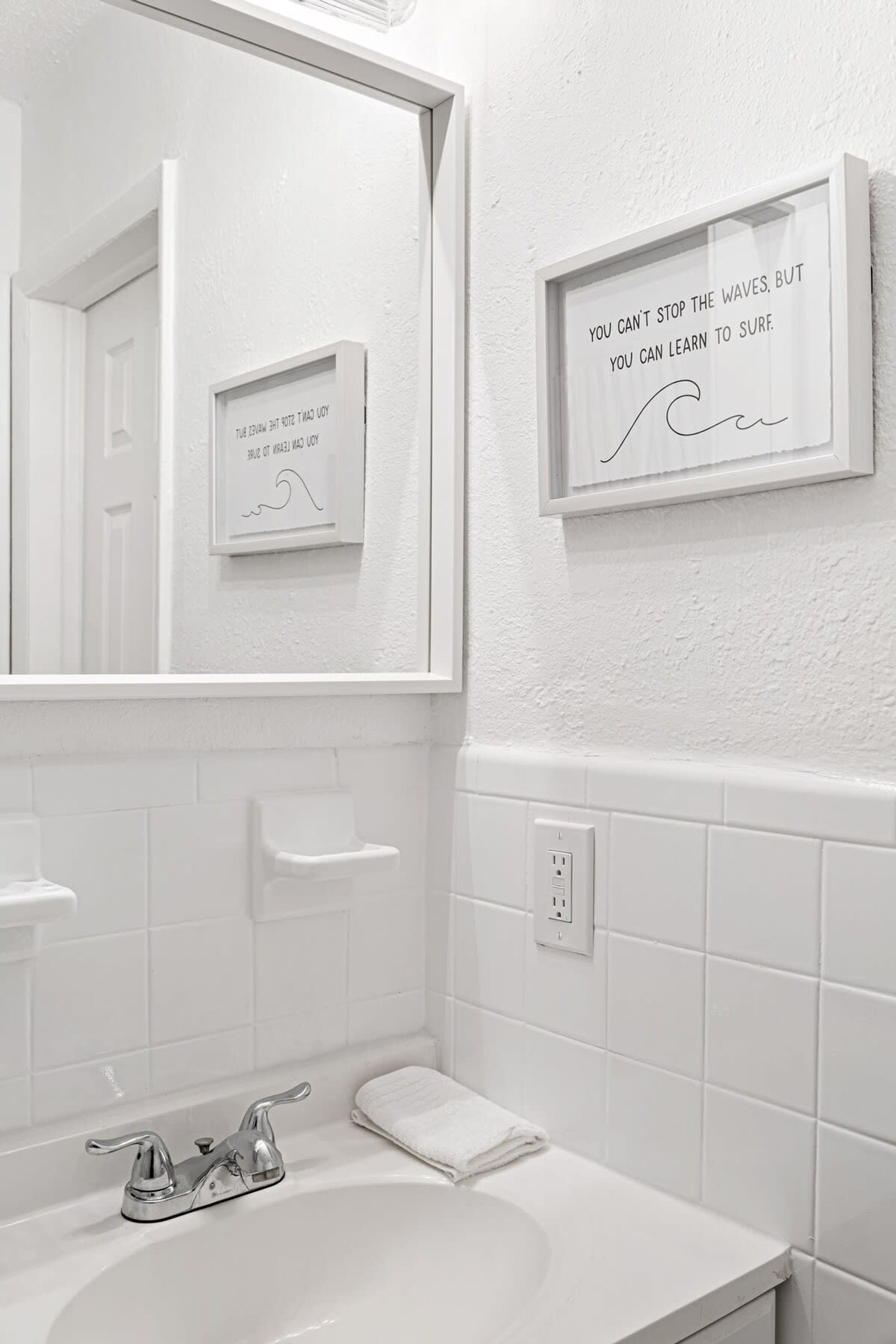 Un baño con un lavamanos blanco, un espejo y dos frases enmarcadas en la pared.