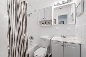 Een moderne badkamer met badkuip, toilet en wastafel. Er zijn witte kastjes en een spiegel boven de wastafel.