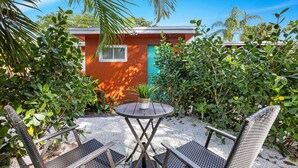 Un patio con una mesa redonda y dos sillas, rodeado de abundante vegetación y un pequeño edificio con fachada roja y azul.