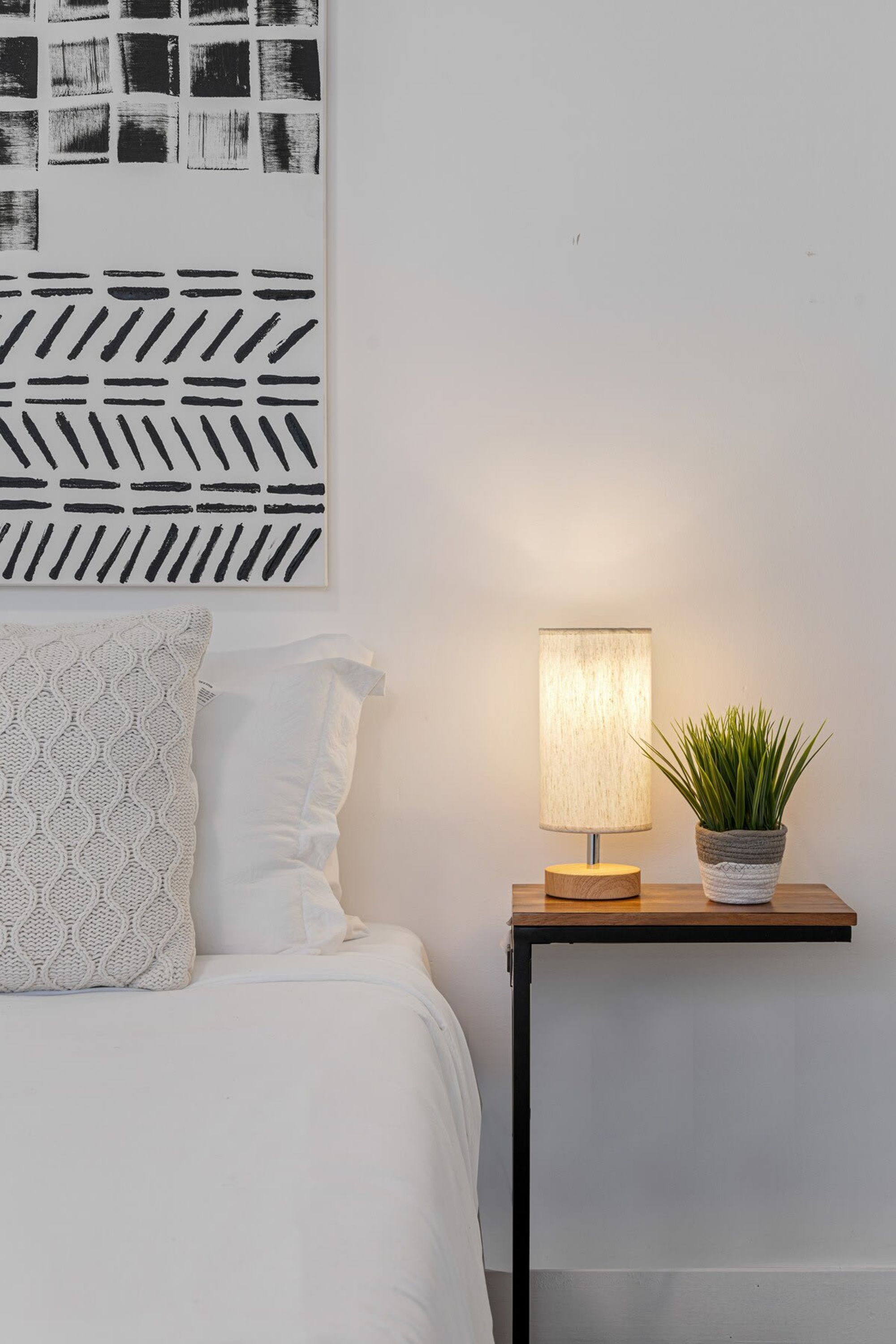 A neatly made bed with white bedding and a patterned pillow, a bedside table with a lamp and a potted plant, and a wall with a geometric black and white artwork.
