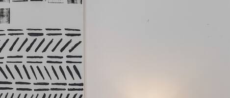 A neatly made bed with white bedding and a patterned pillow, a bedside table with a lamp and a potted plant, and a wall with a geometric black and white artwork.