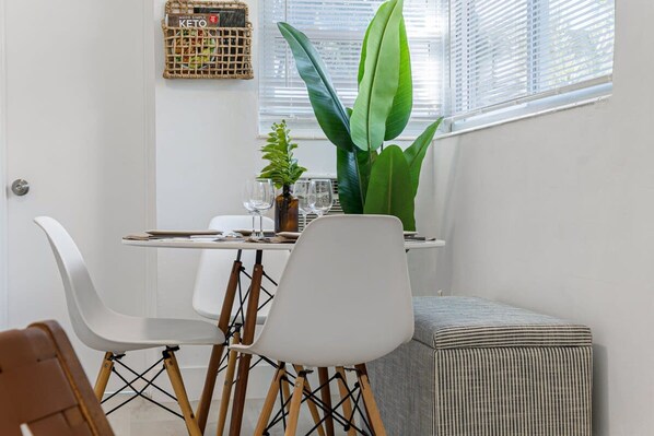 A dining area with a white table, white chairs, and a striped ottoman. There is a large green plant on the windowsill and a woven basket on the wall. - Lake Ridge Villa in Fort Lauderdale (Fort Lauderdale)