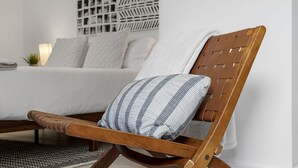 Una silla de madera con un cojín de cuadros en un dormitorio moderno con una cama blanca, una pared estampada y una pequeña planta.