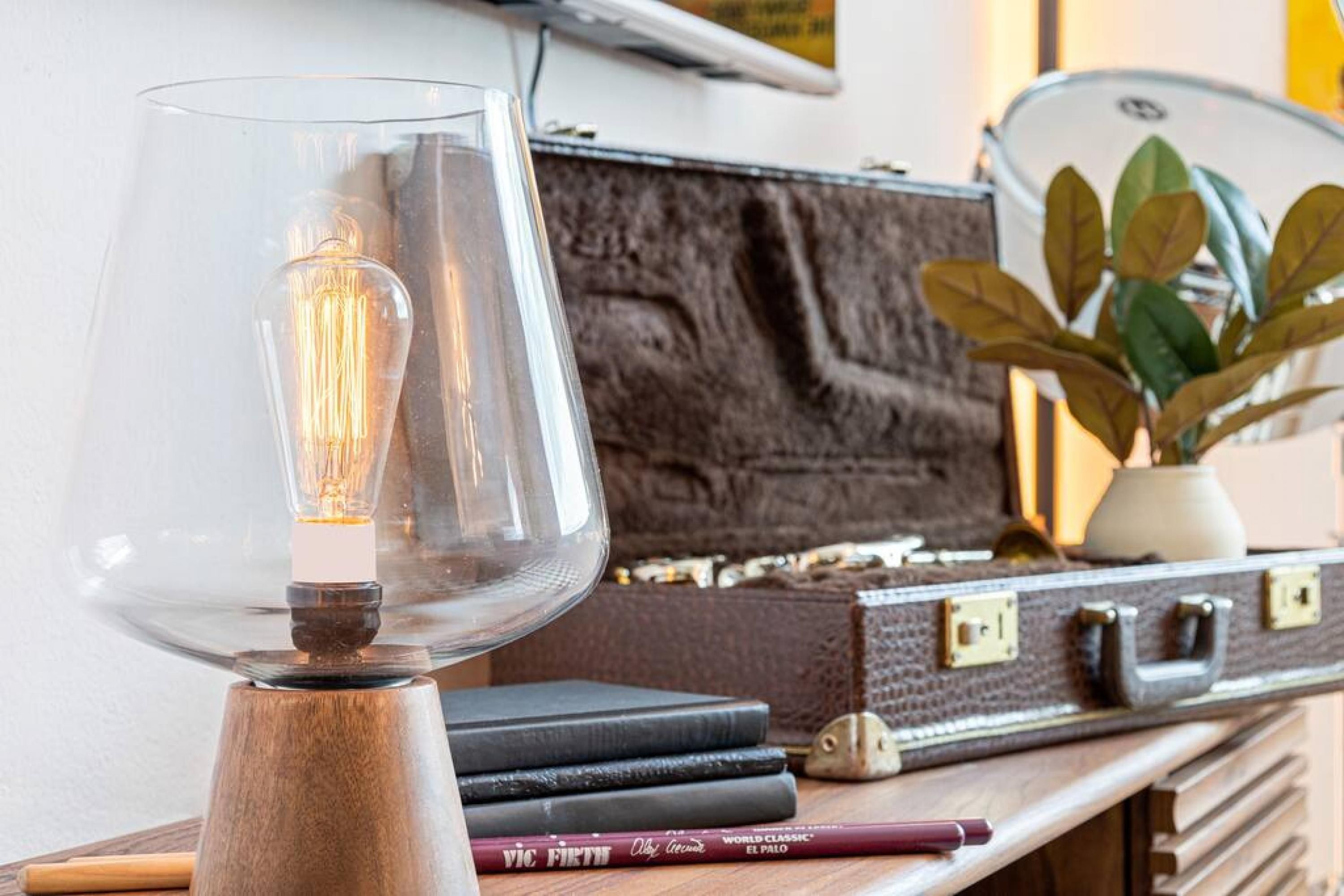 Une lampe dotée d’un abat-jour transparent et d’un pied en bois, posée sur une table avec une valise et des livres.