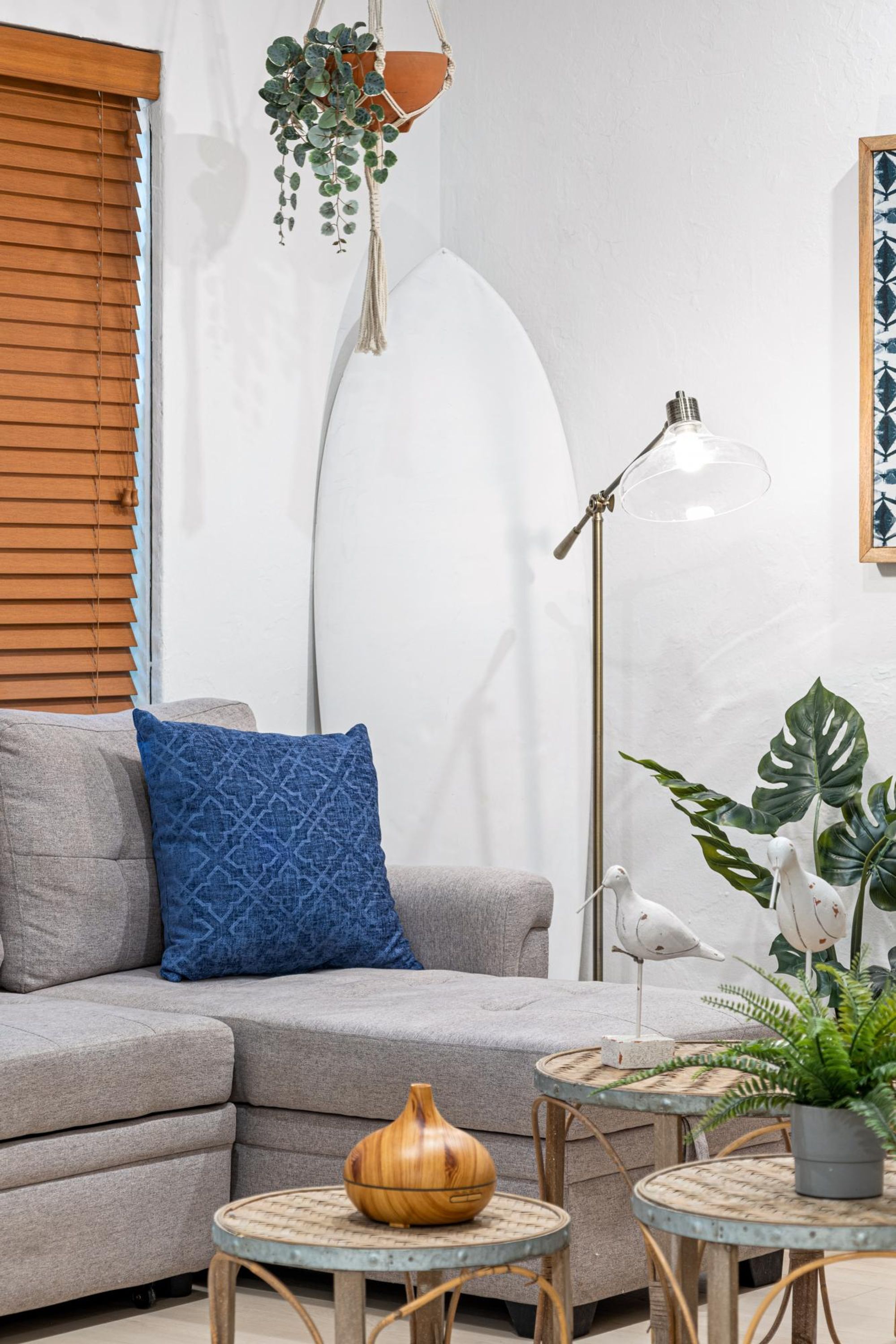 Een woonkamer met een grijze bank, een blauw kussen met patroon, een houten schaal op een klein rond tafeltje en een hangplant.
