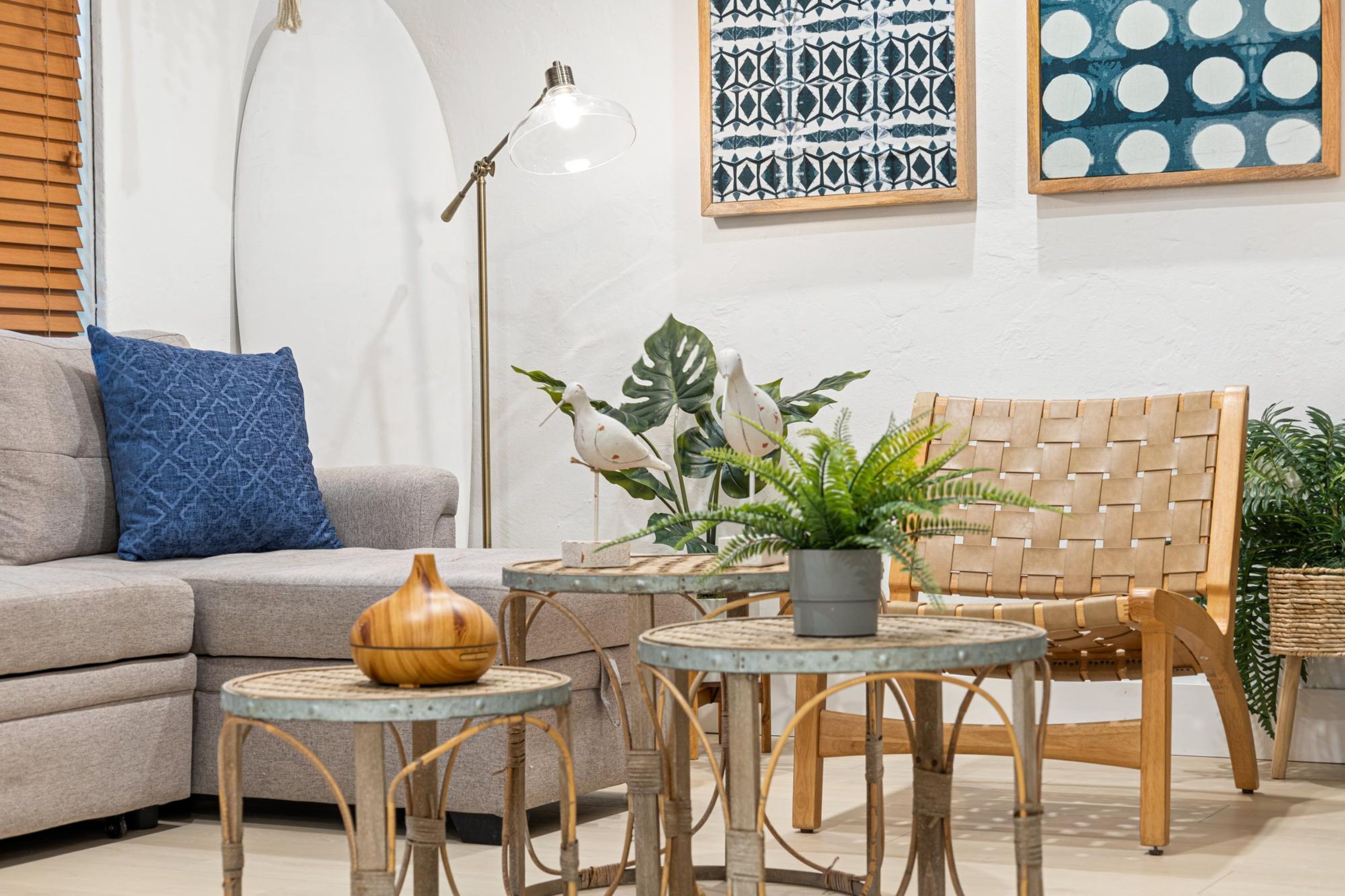A living room with a grey sofa, a blue patterned pillow, a wooden bowl, a woven chair, and two small round tables with plants.