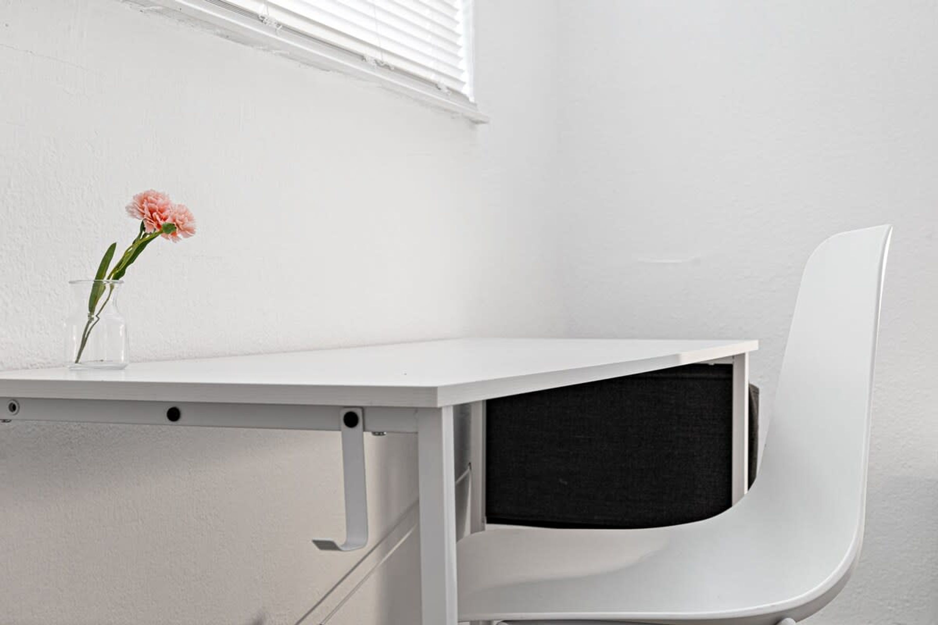 A white desk with a vase containing a pink flower, a white chair, and a window with blinds.