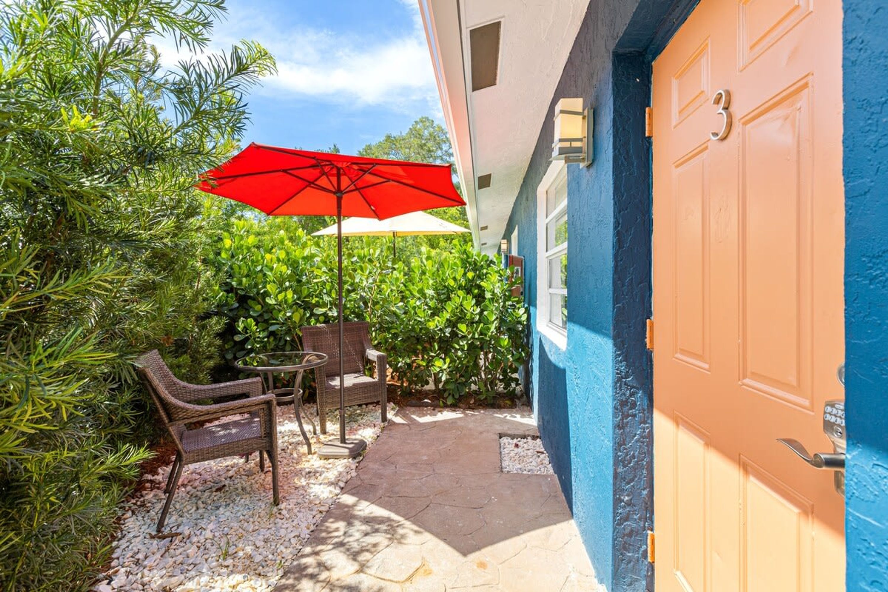 Un patio con mobiliario de mimbre, un paraguas rojo y una puerta azul con el número 3.