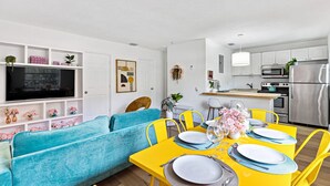 Un salon moderne avec un canapé bleu, une table à manger jaune pour quatre personnes et un coin cuisine doté d’appareils électroménagers en acier inoxydable.