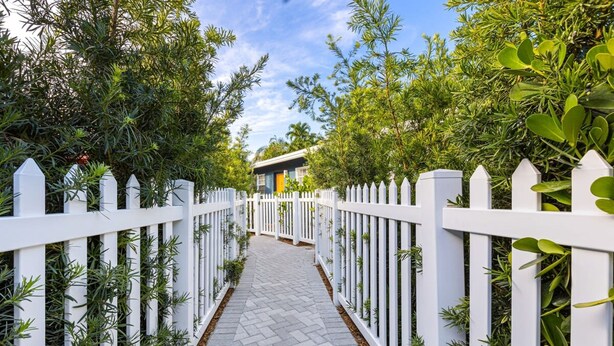 Un sentier bordé de clôtures blanches en bois et d’une végétation luxuriante, qui mène à un bâtiment doté d’une porte jaune.