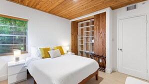 Ein Schlafzimmer mit Holzbalkendecke, einem Bett mit weißer und gelber Bettwäsche, einem Nachttisch, einem Schrank mit Einlegeböden und einem Fenster mit Jalousien.