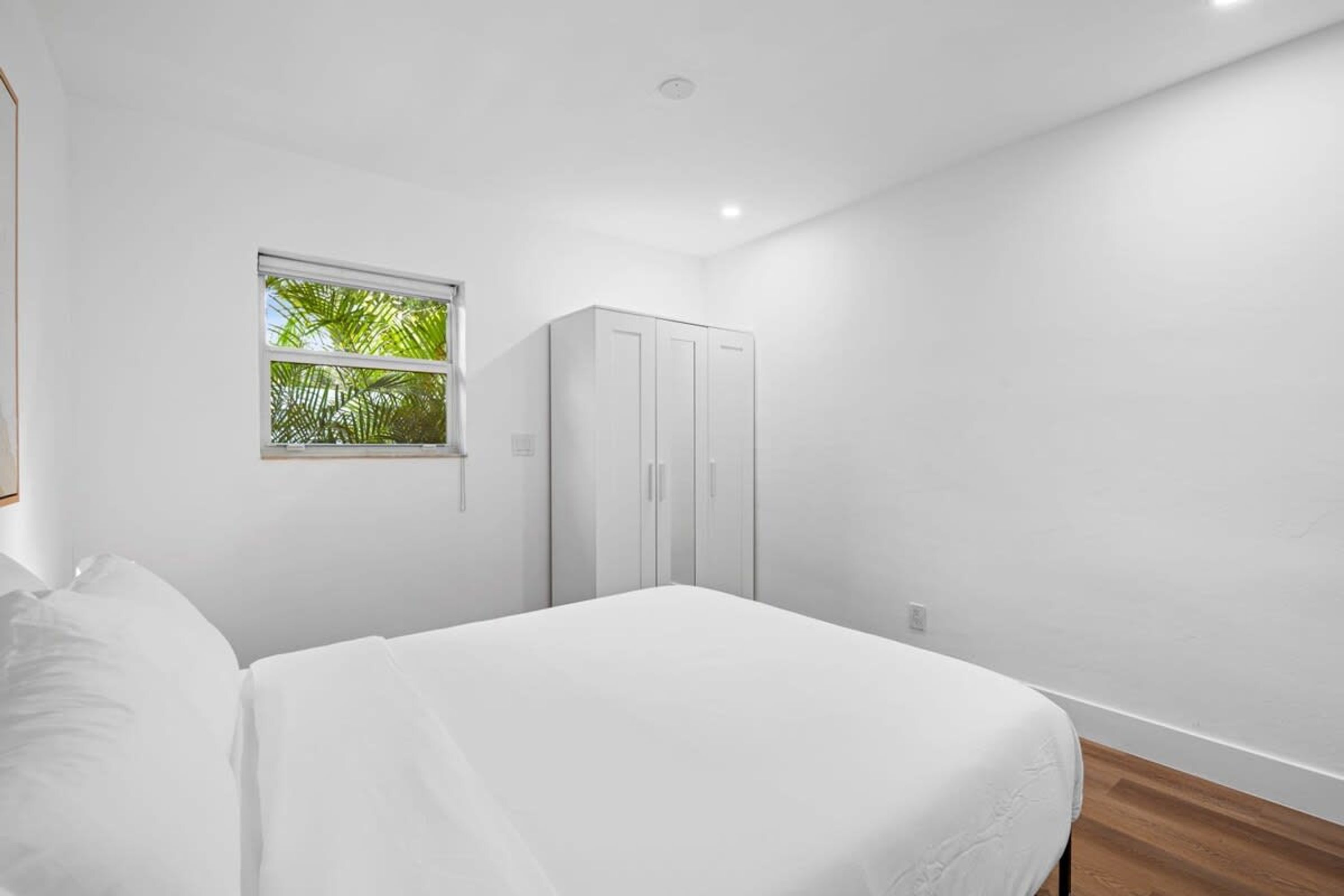 Un dormitorio con cama, armario, ventana con vistas a la vegetación y suelo de madera.