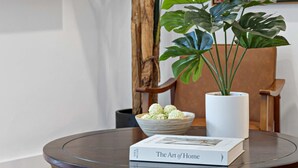 Una mesa redonda con un libro titulado "The Art of Home" y un cuenco de bolas verdes. Una planta grande de hojas anchas está al lado de la mesa.