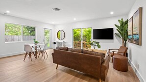 Un salón amplio con un sofá marrón, suelo de madera, un comedor con una mesa y sillas blancas, ventanas grandes con vistas a zonas verdes y un televisor montado en la pared.