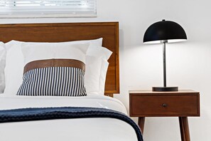 Een keurig opgemaakt bed met witte lakens en gestreepte kussens, een donkerblauwe plaid en een houten nachtkastje met een zwarte lamp ernaast.