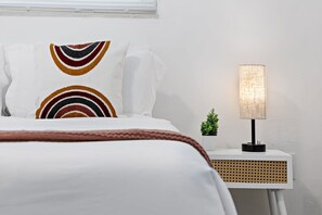 Een net opgemaakt bed met witte lakens en kleurrijke kussens, een bedlampje en een kleine potplant op een witte nachtkastje.