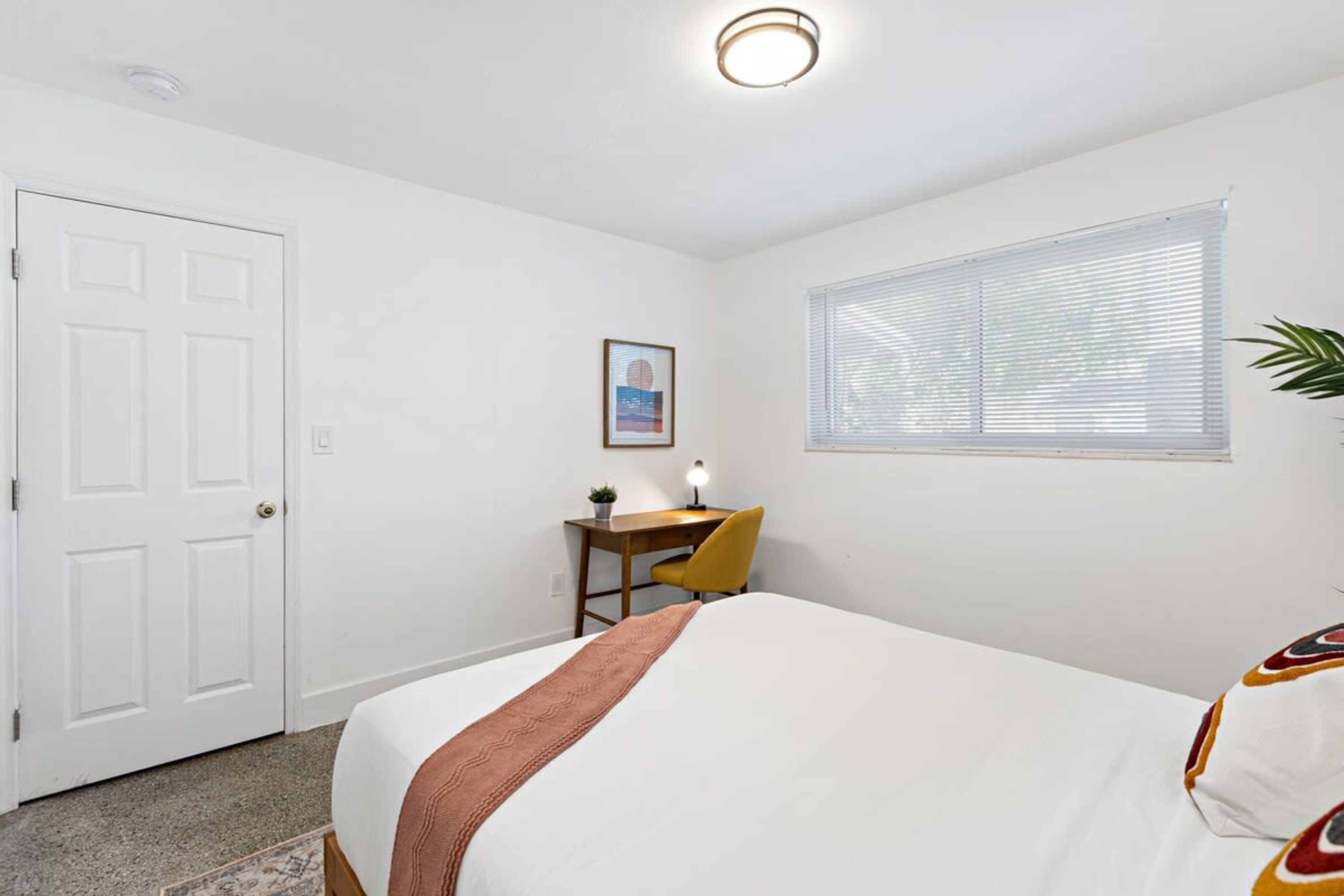 Ein Schlafzimmer mit Bett, Schreibtisch mit Stuhl, Fenster mit Jalousien und einer Tür.