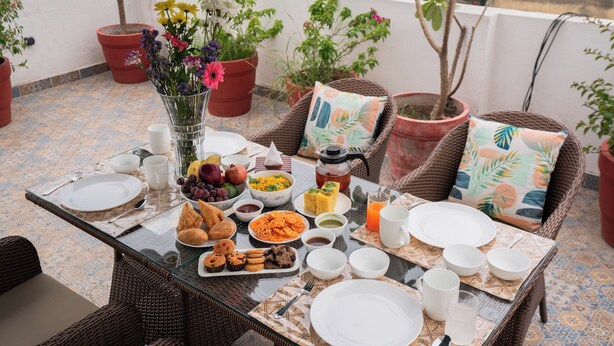 En frukostuppdukning på en balkong, med ett bord dukat för två personer, inklusive tallrikar, koppar och ett urval av olika matvaror.