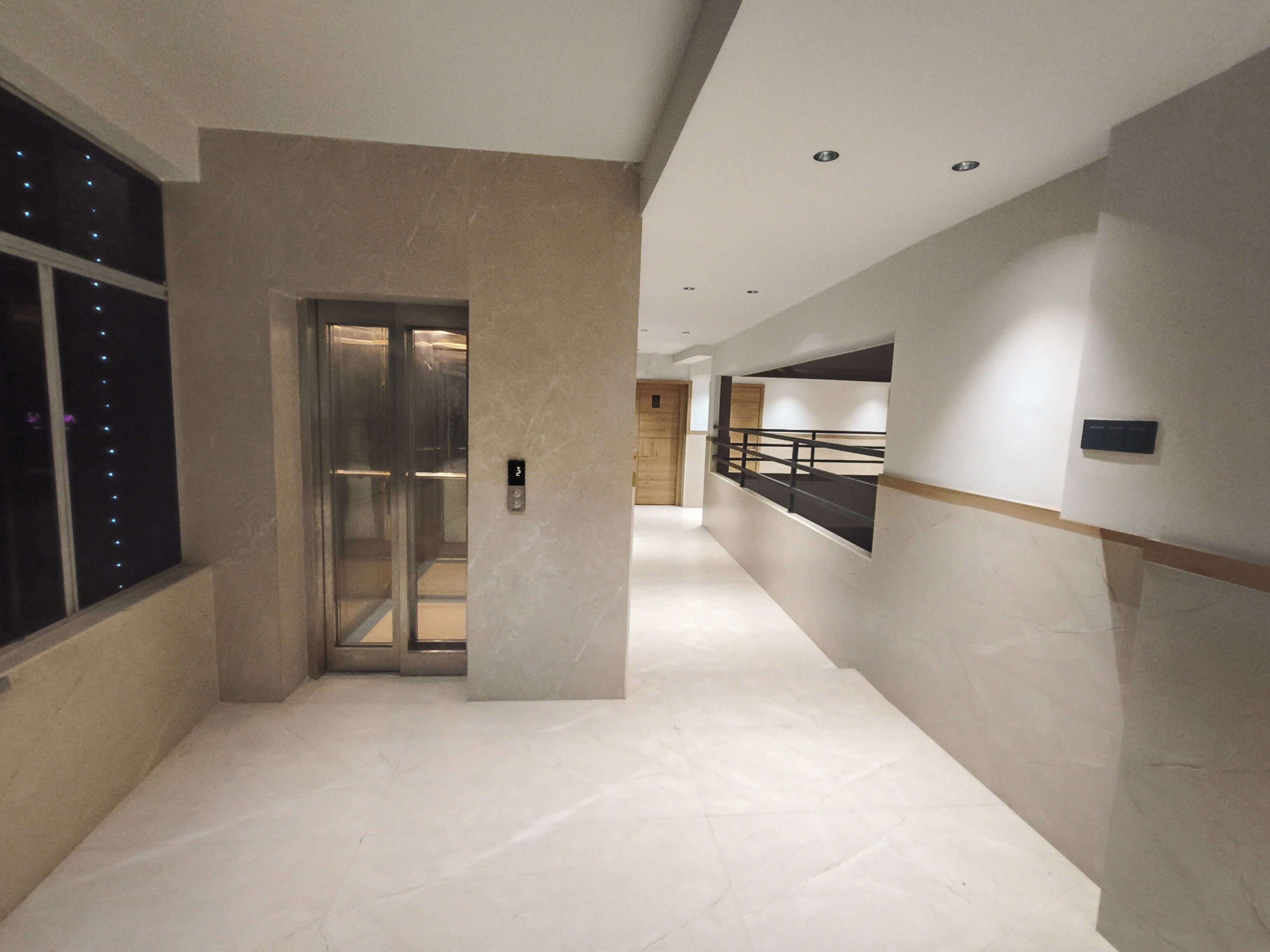 A modern hallway with a glass elevator, a wooden door, and a black railing.