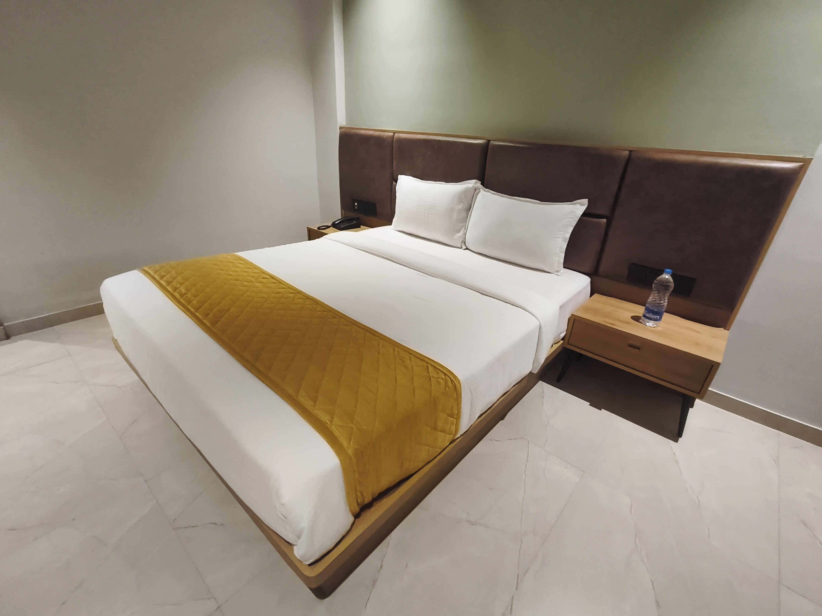 Una habitación de hotel con una cama grande, una mesita de noche con lámpara y una botella de agua en el buró.