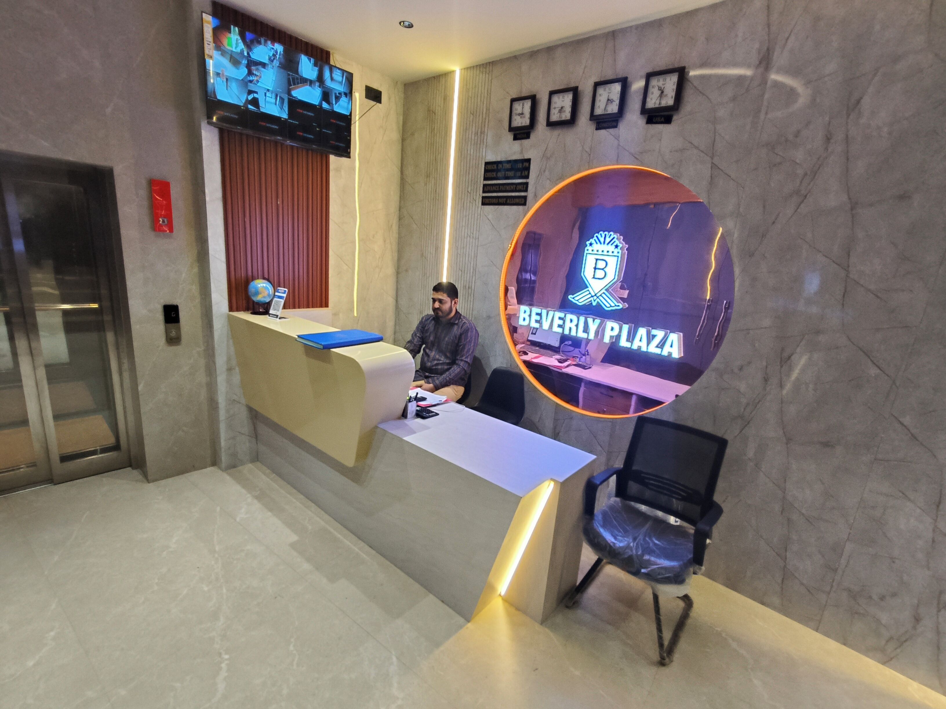A reception desk with a digital screen displaying multiple camera feeds, a clock, and a Beverly Plaza logo.