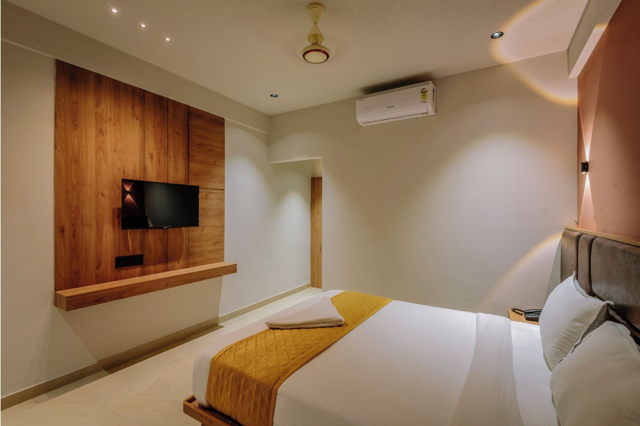 Moderni hotellihuone, jossa on puukoristeinen seinä, sängyn yläpuolelle asennettu litteänäyttöinen televisio, yöpöytä lampun kanssa sekä suuri sänky, jossa on valkoinen peitto ja sinapinkeltainen heittopeitto.