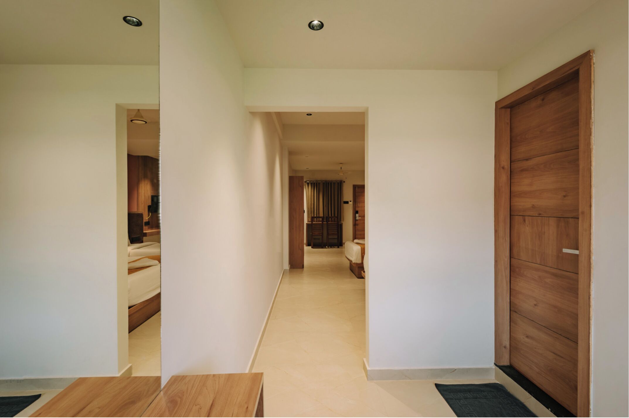 Un pasillo con una puerta de madera a la derecha, una cama a la izquierda y vista a otra habitación.