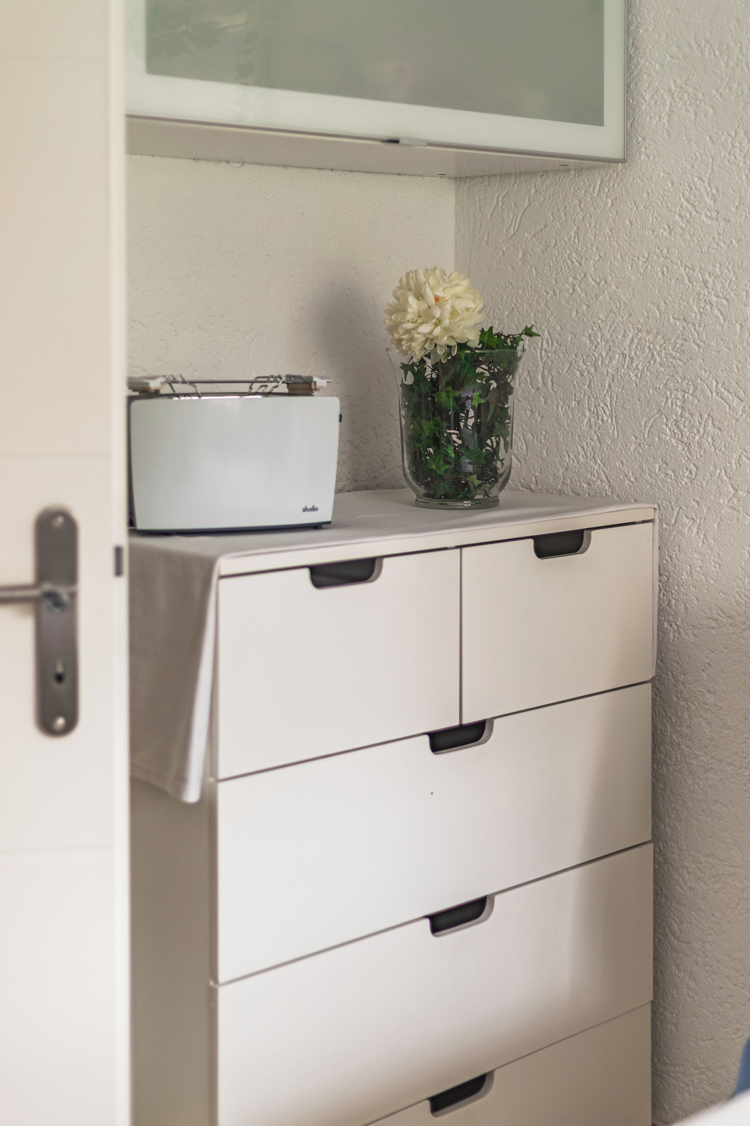 A white cabinet with drawers, a white box on top, and a clear vase with flowers.