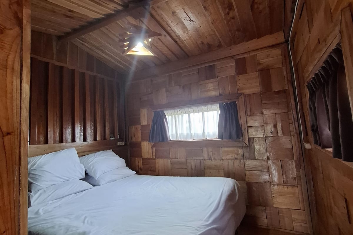 A wooden cabin room with a bed, a window with curtains, and a sloped ceiling.