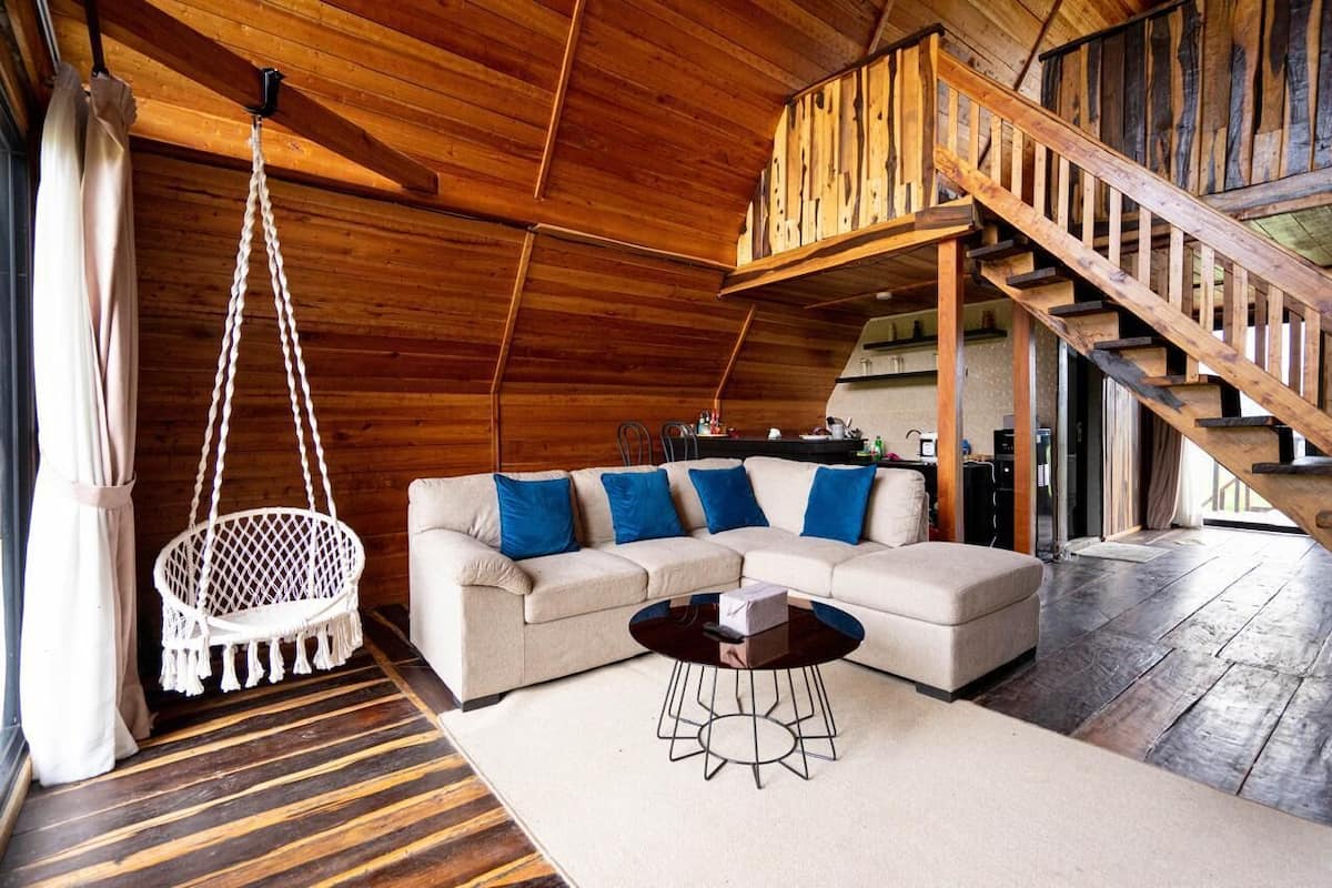 A cozy living room with a wooden ceiling, a hanging chair, a sectional sofa with blue cushions, and a staircase leading to an upper level.