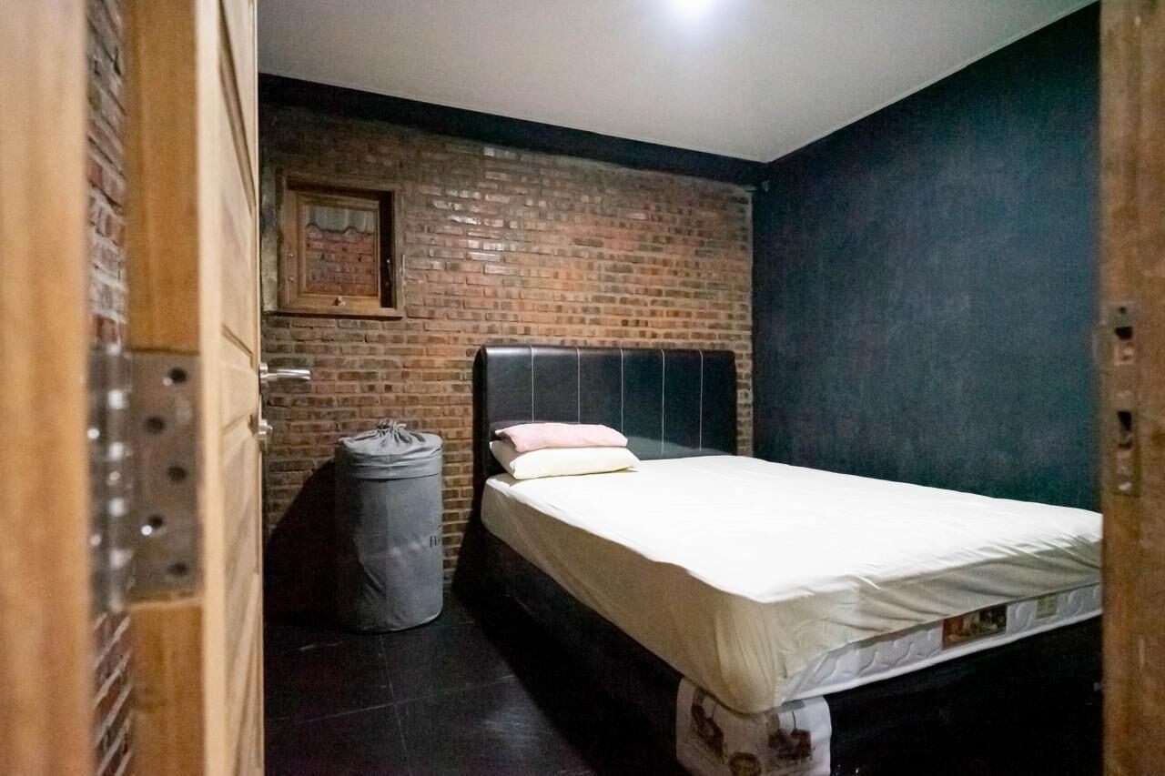 Une petite pièce, de taille modeste, avec un lit simple, un mur en briques et une porte de couleur sombre.