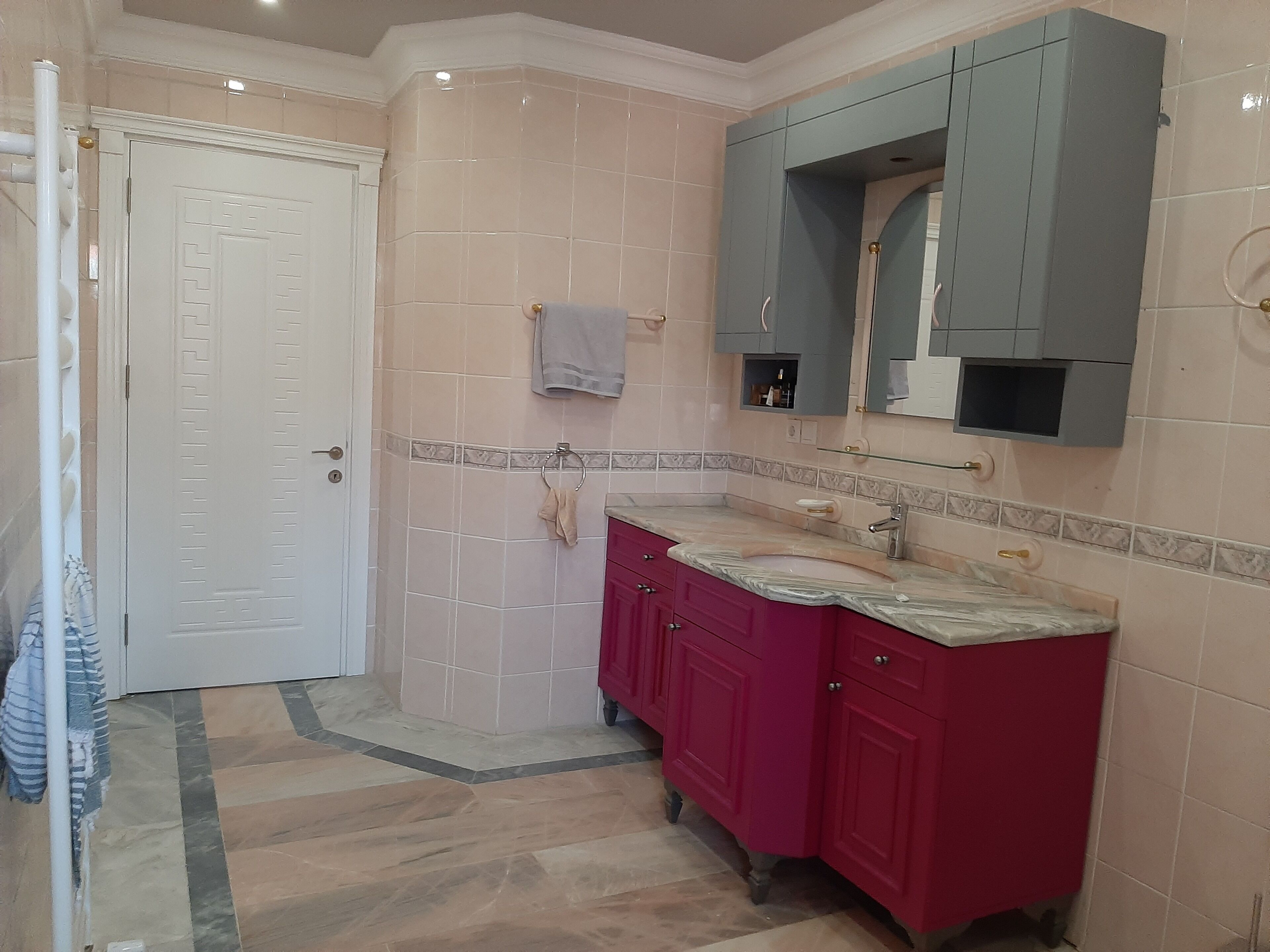 Un baño con puerta blanca, paredes de azulejos rosa, un lavabo de mármol y un espejo sobre el lavabo.